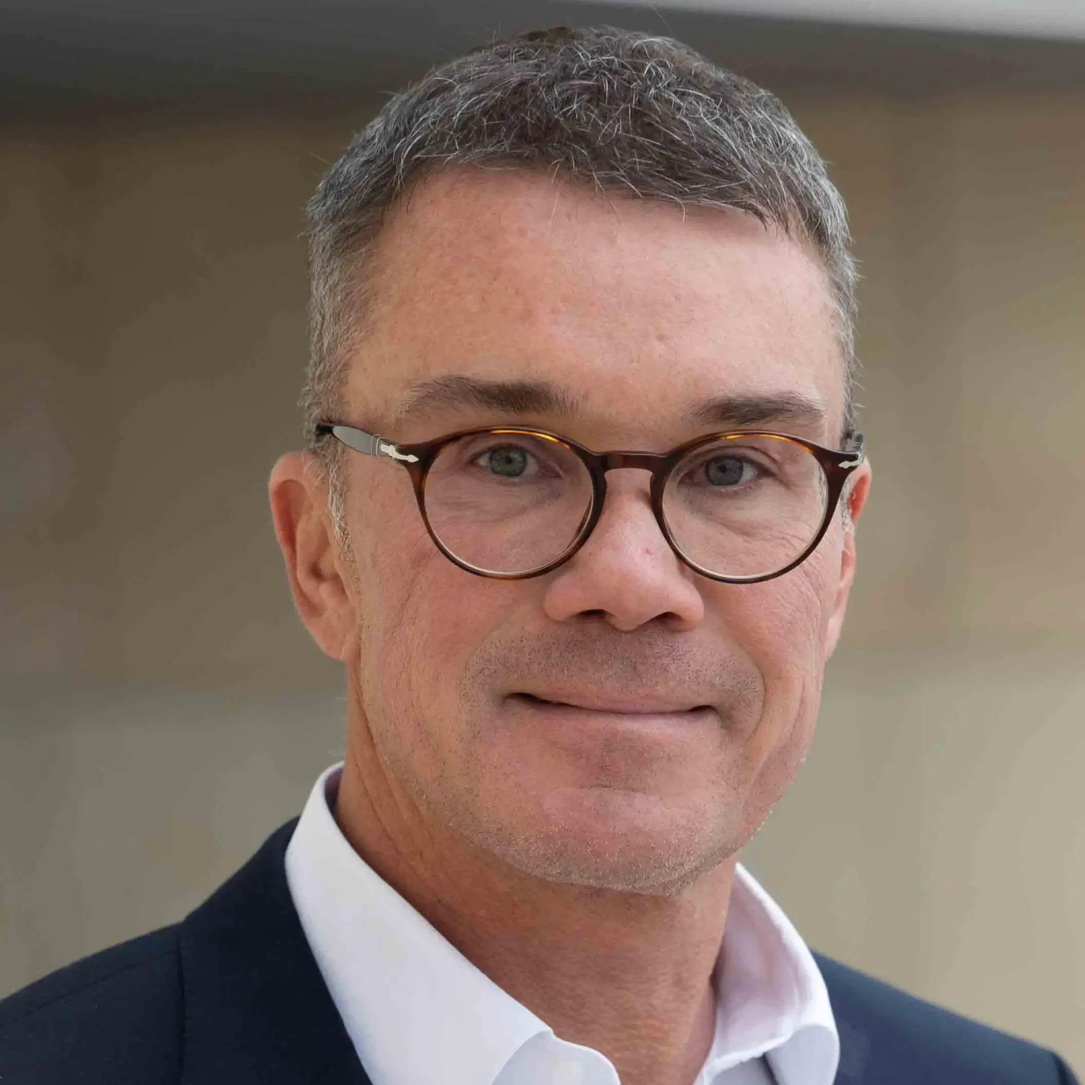 A middle-aged man with short gray hair, wearing round eyeglasses, a white collared shirt, and a dark suit jacket, smiling slightly in front of a blurred neutral background.