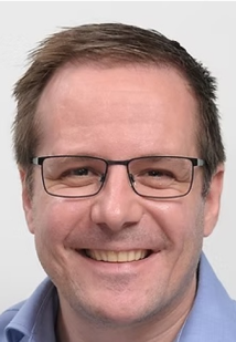 A man with short brown hair and glasses smiles at the camera. He is wearing a light blue collared shirt and is set against a plain, light background.