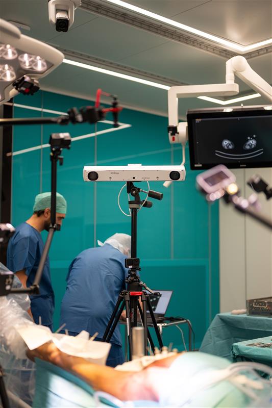 Two medical professionals in scrubs work in a brightly lit operating room, surrounded by surgical equipment, cameras, monitors, and advanced imaging devices. A patient lies on the operating table in the foreground.