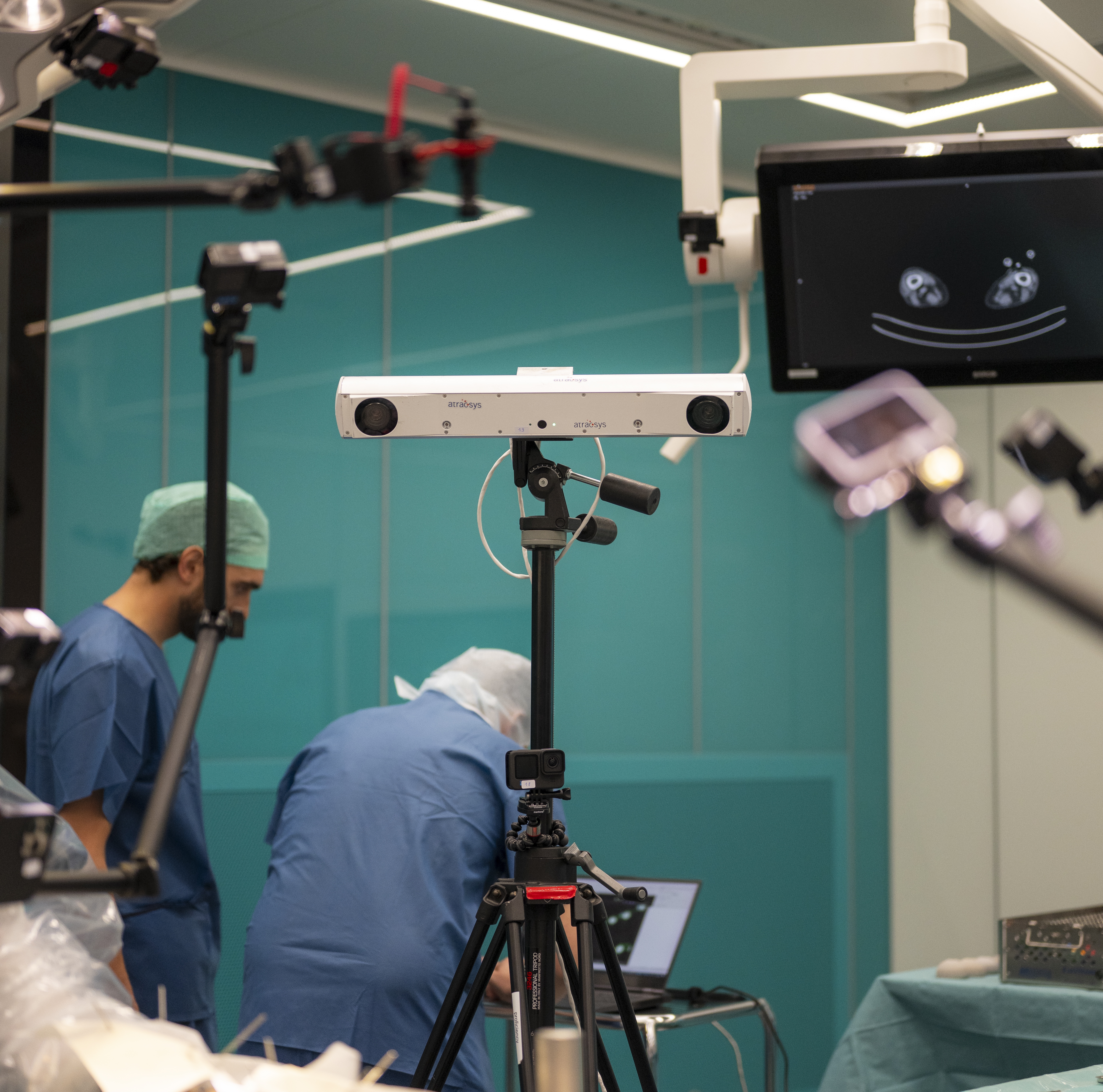 A medical imaging device on a tripod stands in an operating room, with two medical professionals in scrubs and caps working nearby. A monitor displays medical images in the background.